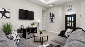 A modern living room with a grey sofa set, a round coffee table, and a flat-screen TV mounted on the wall.