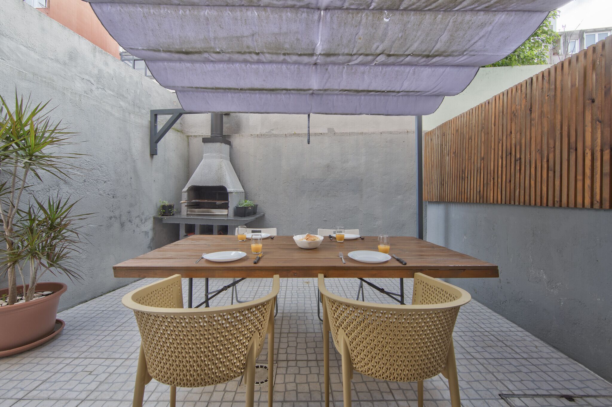 An outdoor dining area with a wooden table, two wicker chairs, a grill, and a potted plant.