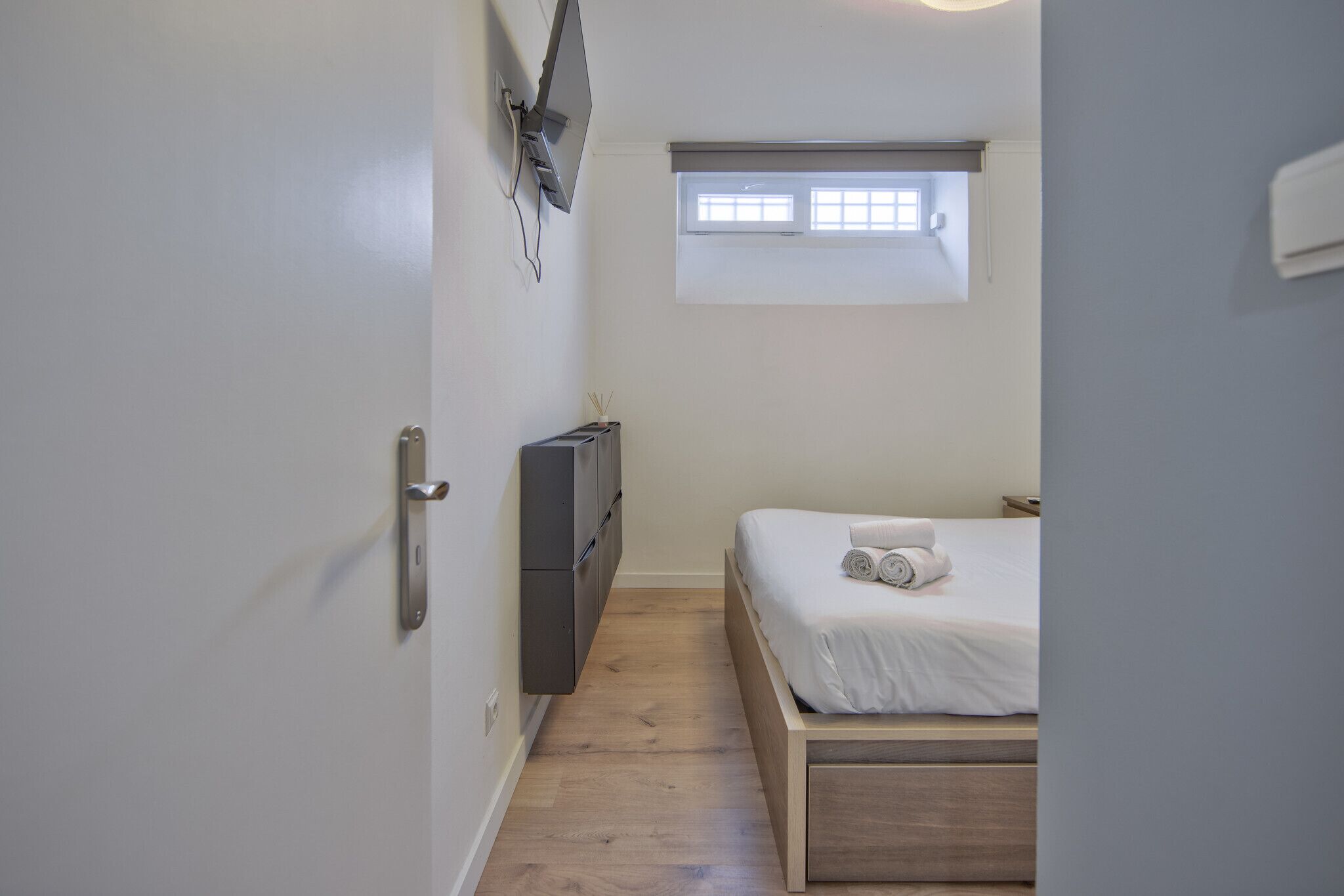 A small, neatly arranged bedroom with a single bed, a window with blinds, a flat-screen TV mounted on the wall, and a small cabinet.