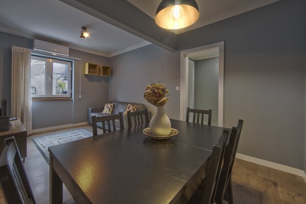 A dining area with a wooden table, chairs, a vase with flowers, a window with curtains, and a ceiling light fixture.