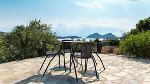 Une terrasse extérieure avec un sol en pierre et des chaises en osier, offrant une vue panoramique sur un paysage magnifique, avec des montagnes et une étendue d’eau.