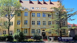 A yellow building with multiple windows and a red-tiled roof, surrounded by trees and a sign indicating local information. - Rentumi Hostel Świnoujście Noclegi (Swinoujscie)