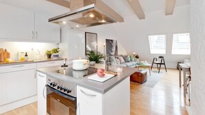 A modern kitchen with white cabinets, a central island, and a built-in oven. There is a dining area with a sofa and a coffee table. - Urban Apartment - City Center (Bergen)