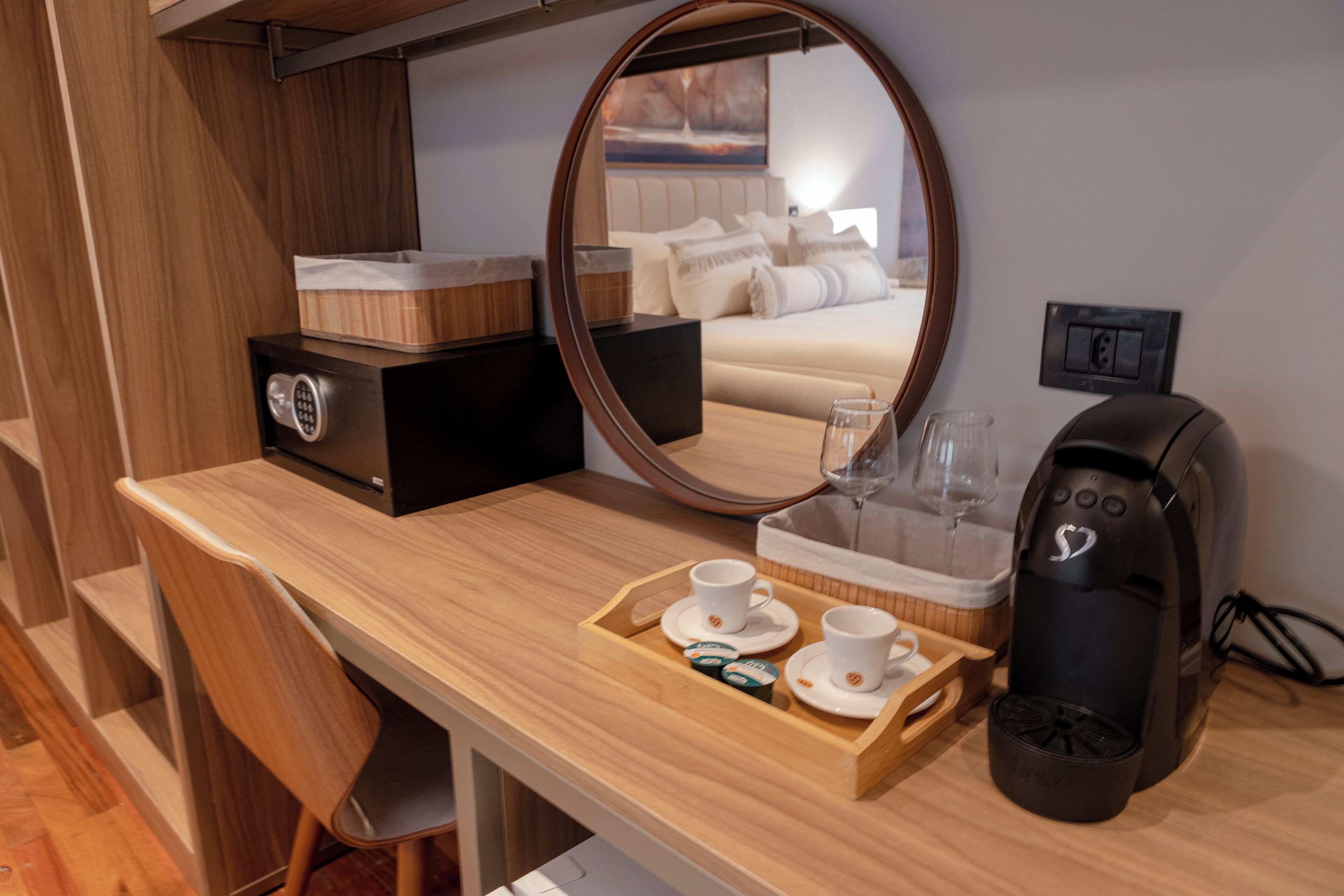 Habitación de hotel con un escritorio de madera, cafetera, bandeja con tazas y un vaso, y una cama con vistas al exterior.