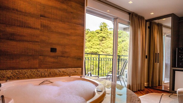 A modern bathroom with a large bathtub, a glass-enclosed shower area, and a view of a balcony with outdoor furniture. - Champet Boutique Hotel (Campos do Jordão)