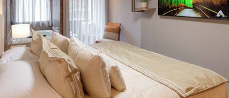 A hotel room with a large bed, a TV on the wall, a desk with a lamp, and a window with curtains.