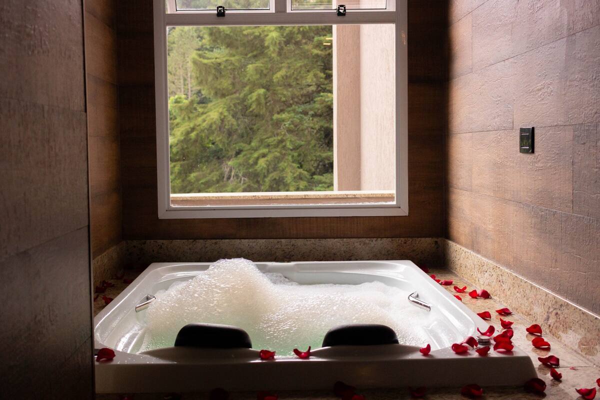 A bathtub with bubbles and rose petals, a window with a forest view, and a stone wall.