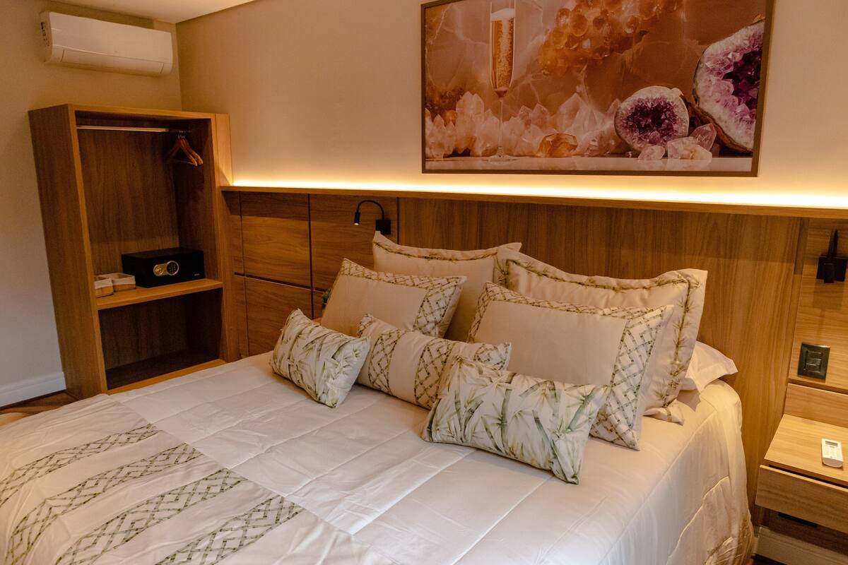 A neatly made bed with floral patterned pillows, a wooden wardrobe, and a wall-mounted artwork depicting fruits and a glass.