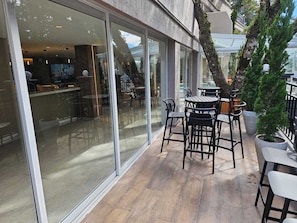 An outdoor seating area with tables and chairs, a glass wall, and a potted tree.