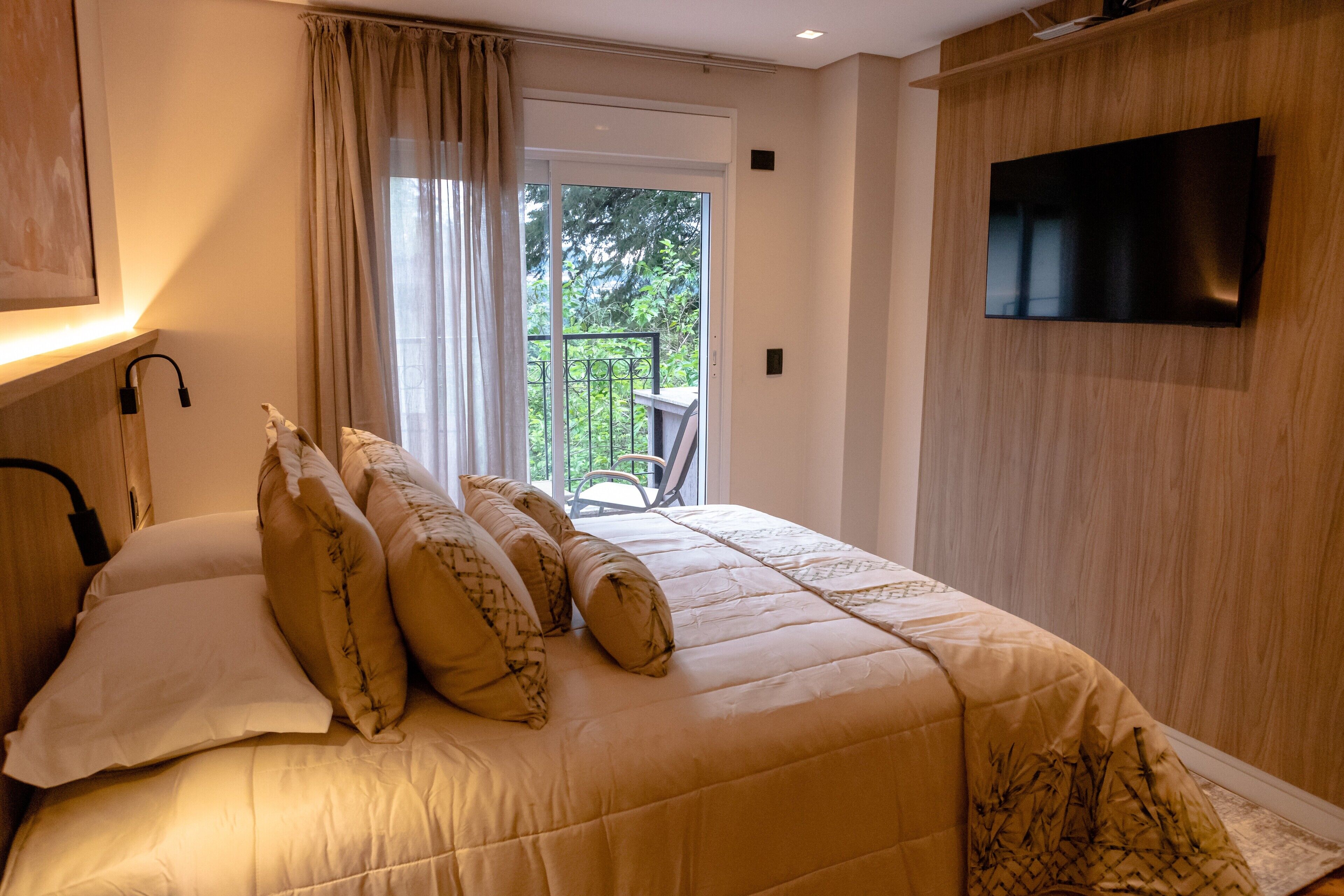 Un dormitorio con cama, televisor montado en la pared y balcón con una silla.