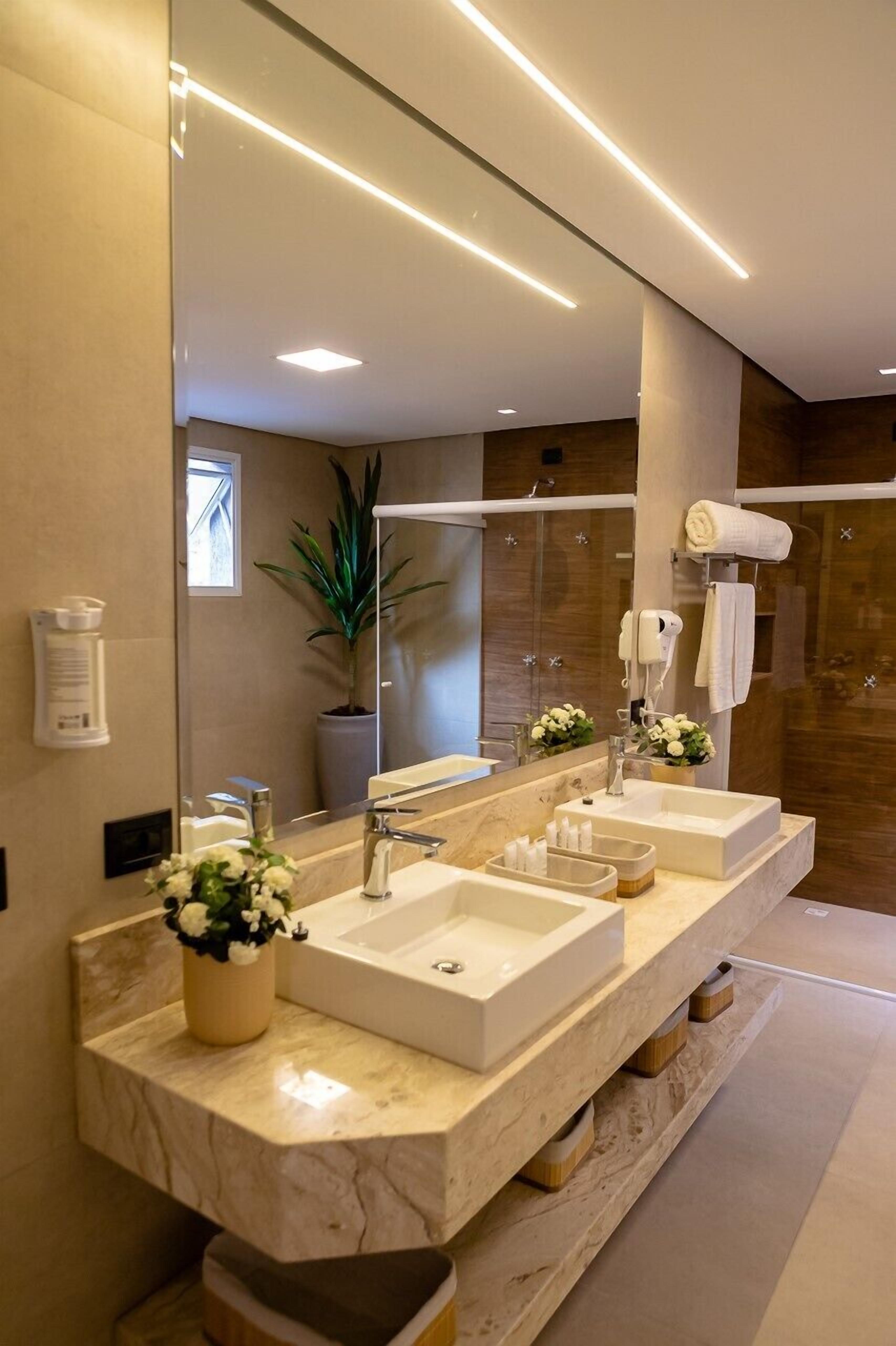 A modern bathroom with a large mirror, two sinks, a potted plant, and a shower area.