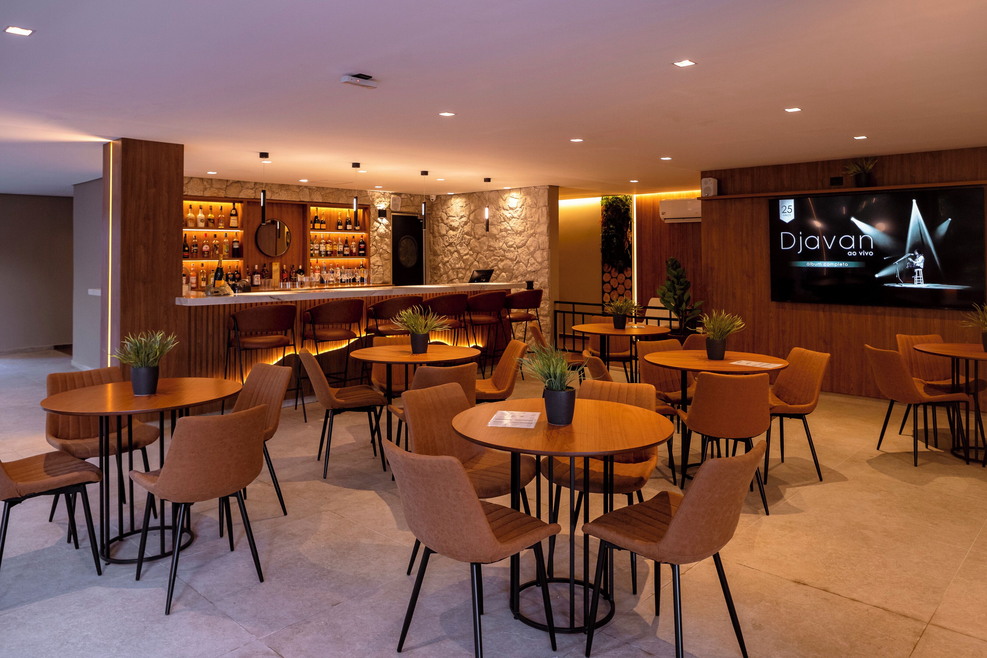 Uma área de bar com uma televisão exibindo 'Djavan do vivo', várias mesas com cadeiras e um balcão repleto de garrafas.