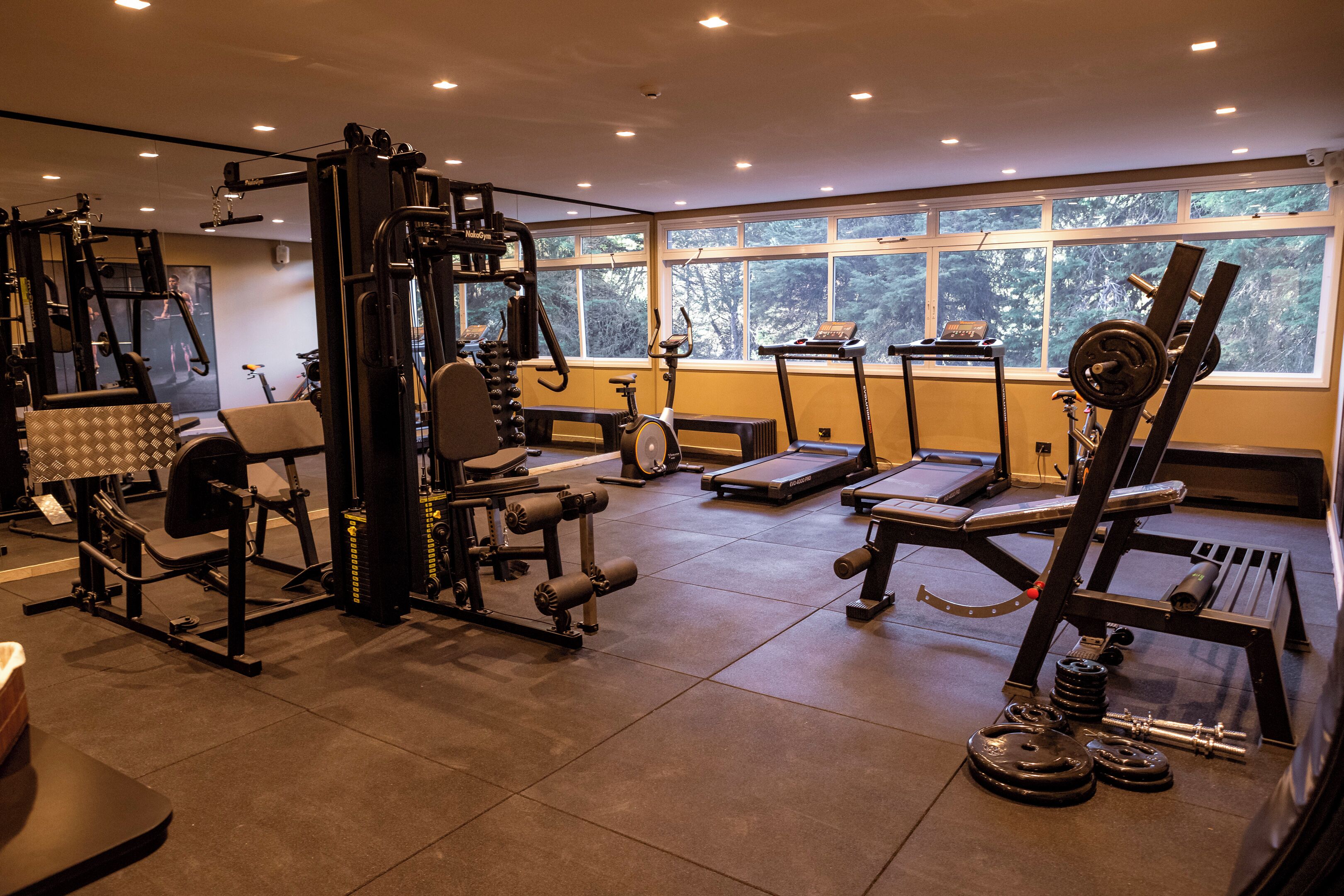 Un gimnasio con diferentes equipos de ejercicio, amplias ventanas y piso de baldosas.