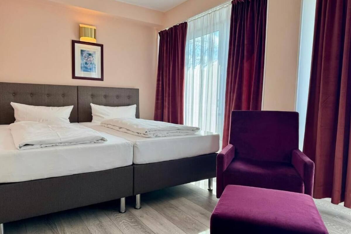 A hotel room with two beds, a large window with curtains, a framed picture on the wall, and a purple armchair.