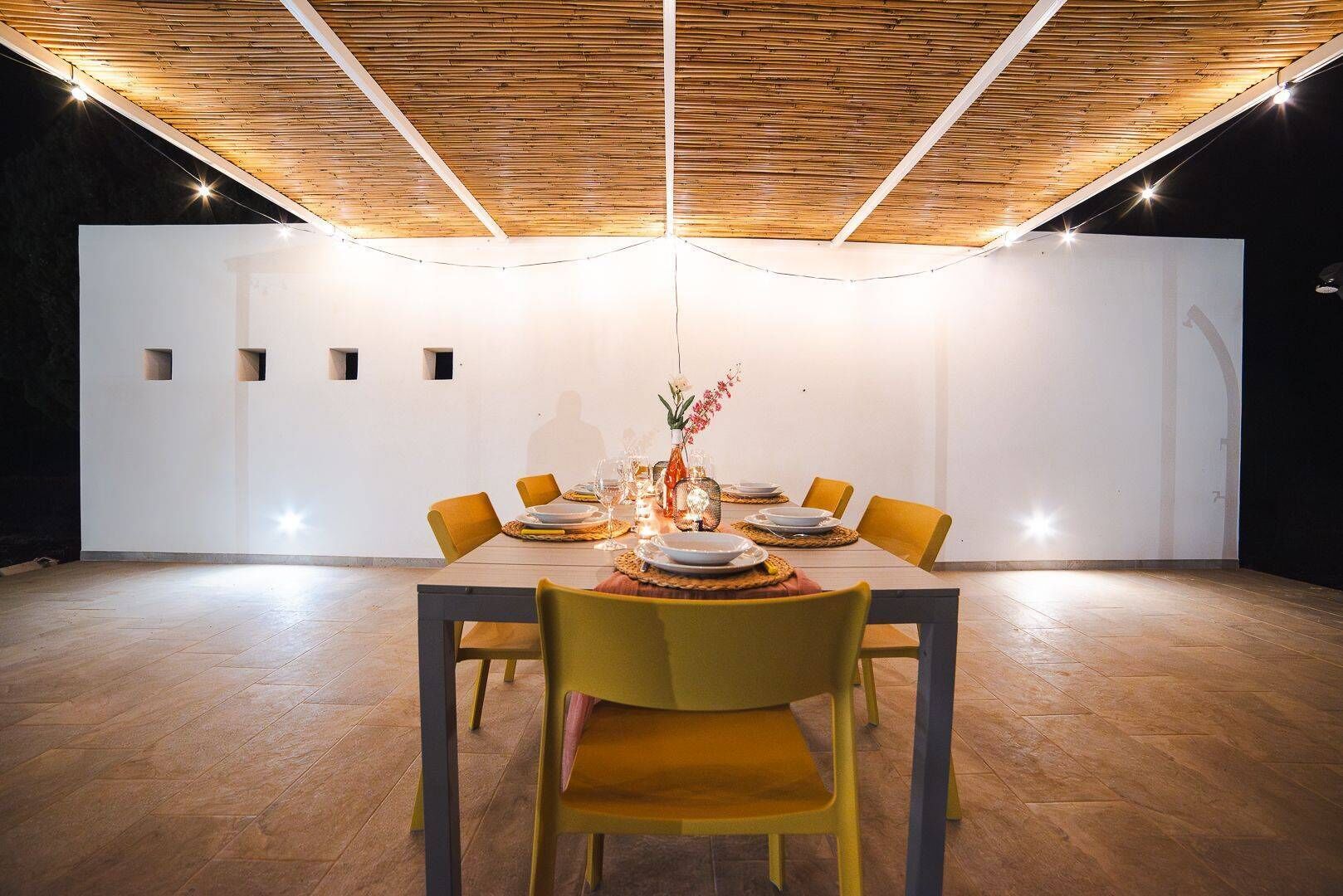 Ein Essbereich mit Holzbalkendecke, einer weißen Wand mit kleinen Fenstern und einem für vier Personen gedeckten Tisch mit gelben Stühlen.