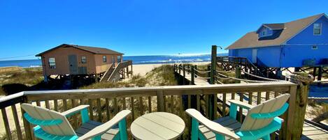 A wooden deck with turquoise chairs and a small round table, overlooking a beach with a wooden structure and a blue building.
