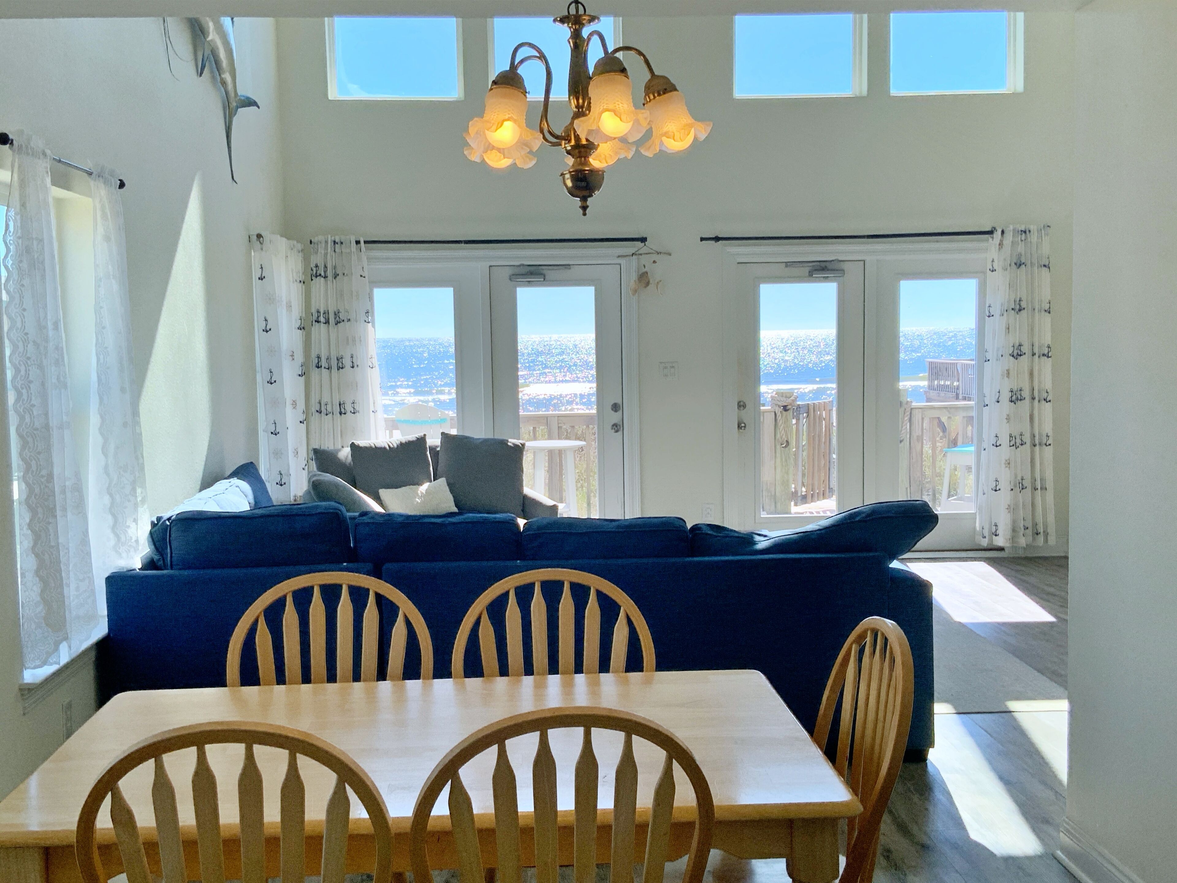 Ein Wohnzimmer mit blauem Sofa, Holzstühlen, einem Esstisch und einer Deckenleuchte. Große Fenster mit Vorhängen bieten Blick auf die Stadt.