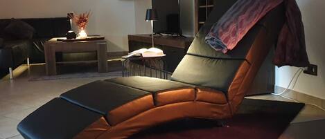 A modern living room with a reclining chair, a flat-screen TV, a small table with a lamp, and a bookshelf.