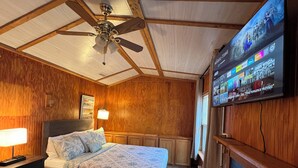 A bedroom with a bed, wooden walls, a ceiling fan, a TV mounted on the wall, and a nightstand with a lamp. - Peaceful Entire 4 BR 2 BA Cabin with AC, WiFi, Pond  View, Recreational room (Crisfield)