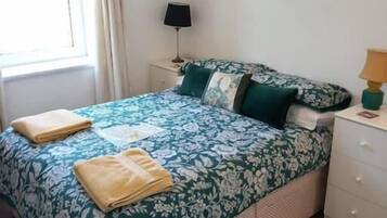 A neatly arranged bedroom with a floral bedspread, a bedside table with a lamp, and a small table with snacks and drinks.