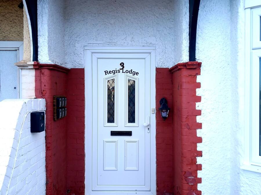 Una puerta blanca con el número 3 y el nombre "Regis Lodge" en la parte superior, flanqueada por muros de ladrillo rojo y un muro de ladrillo blanco.