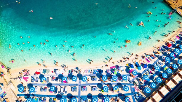An aerial view of a beach with rows of sunbeds and umbrellas, clear turquoise water, and a pier extending into the sea. - Sea View Apartment Terazza Grande (Crikvenica)