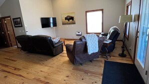 Un salon spacieux avec un plancher en bois, une grande fenêtre, un téléviseur à écran plat, un canapé, un fauteuil et une lampe.