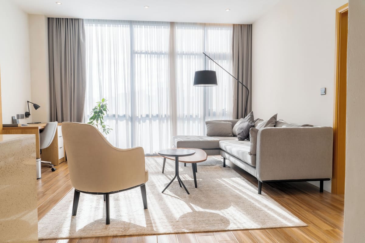 Una sala de estar moderna con sofá, sillón y mesa de centro, ventanas amplias con cortinas y un escritorio con lámpara.