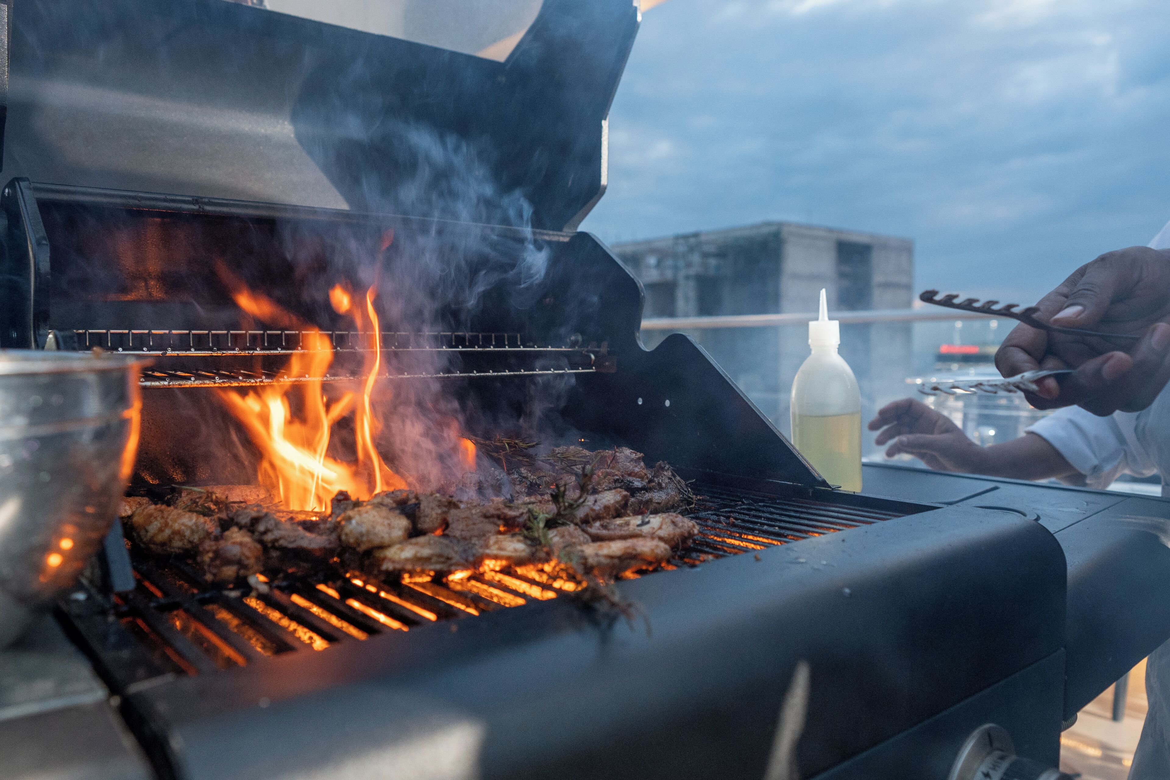 A grill with meat being cooked, a bottle of sauce, and a person grilling.