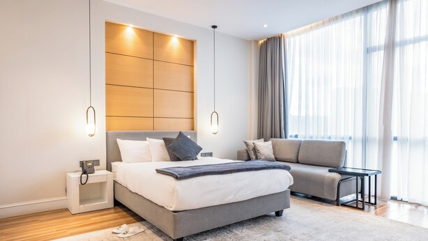 A modern bedroom with a large bed, a sofa, a bedside table with a phone, and a large window with curtains. - Loft Hotel Apartment (Addis Ababa)