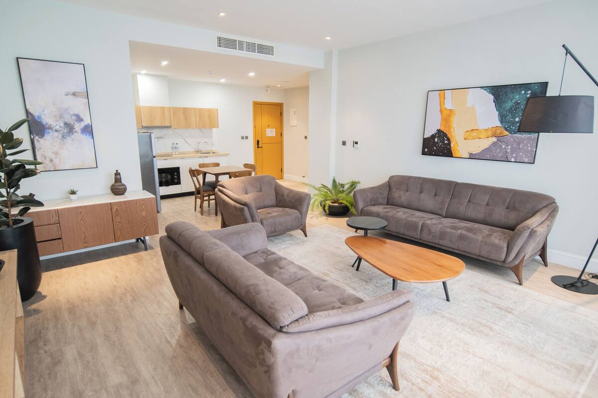 Una sala de estar moderna con un sofá, sillones y una mesa de centro. Se observa una cocina al fondo con armarios y un refrigerador.