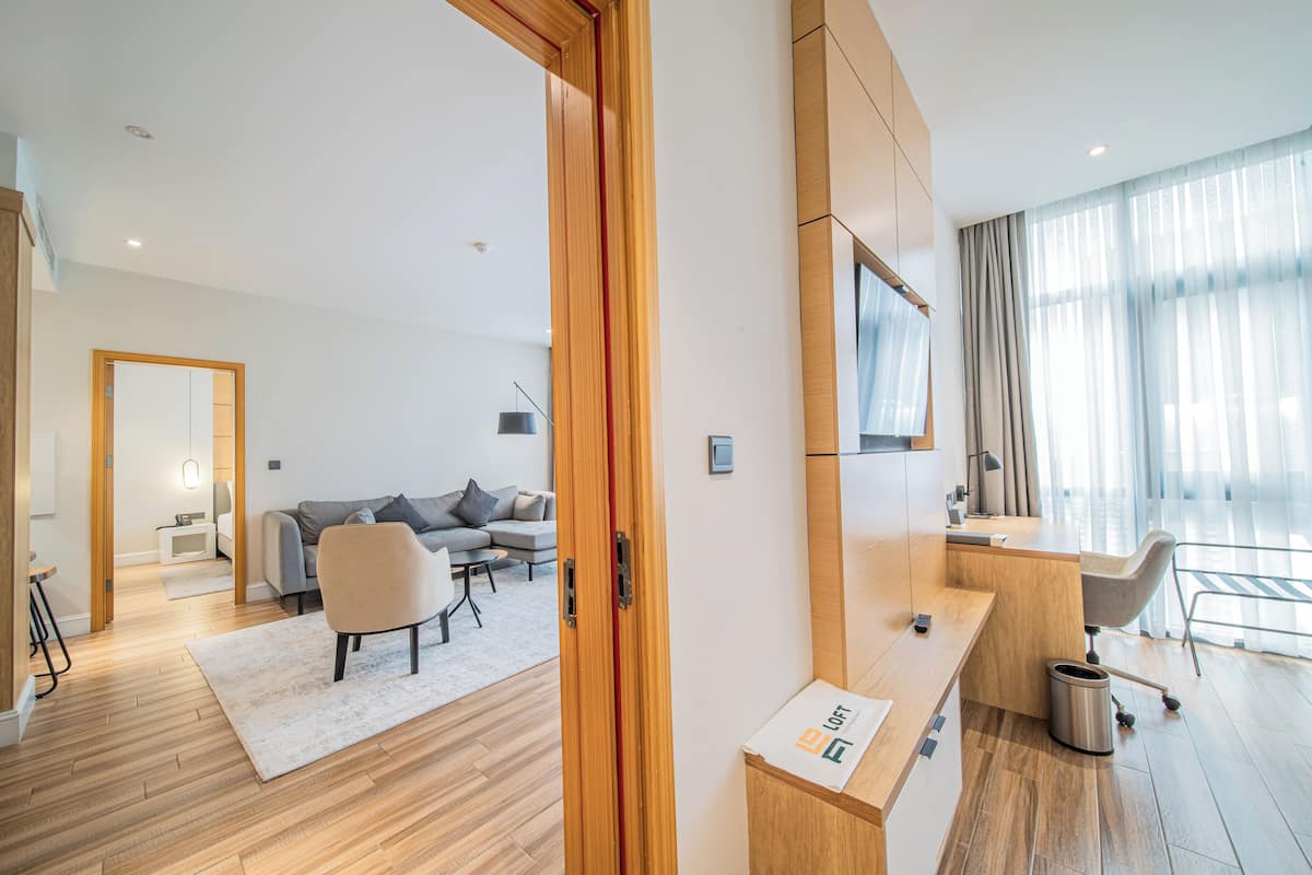 Una habitación de hotel moderna con piso de madera, un sofá gris, un escritorio de madera y un ventanal con cortinas translúcidas.