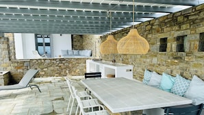 An outdoor patio area with stone walls, a long table, white chairs, and hanging wicker lamps. - Villa louise, sea view, swimming pool, sleeps 12, near Drios (Drios)