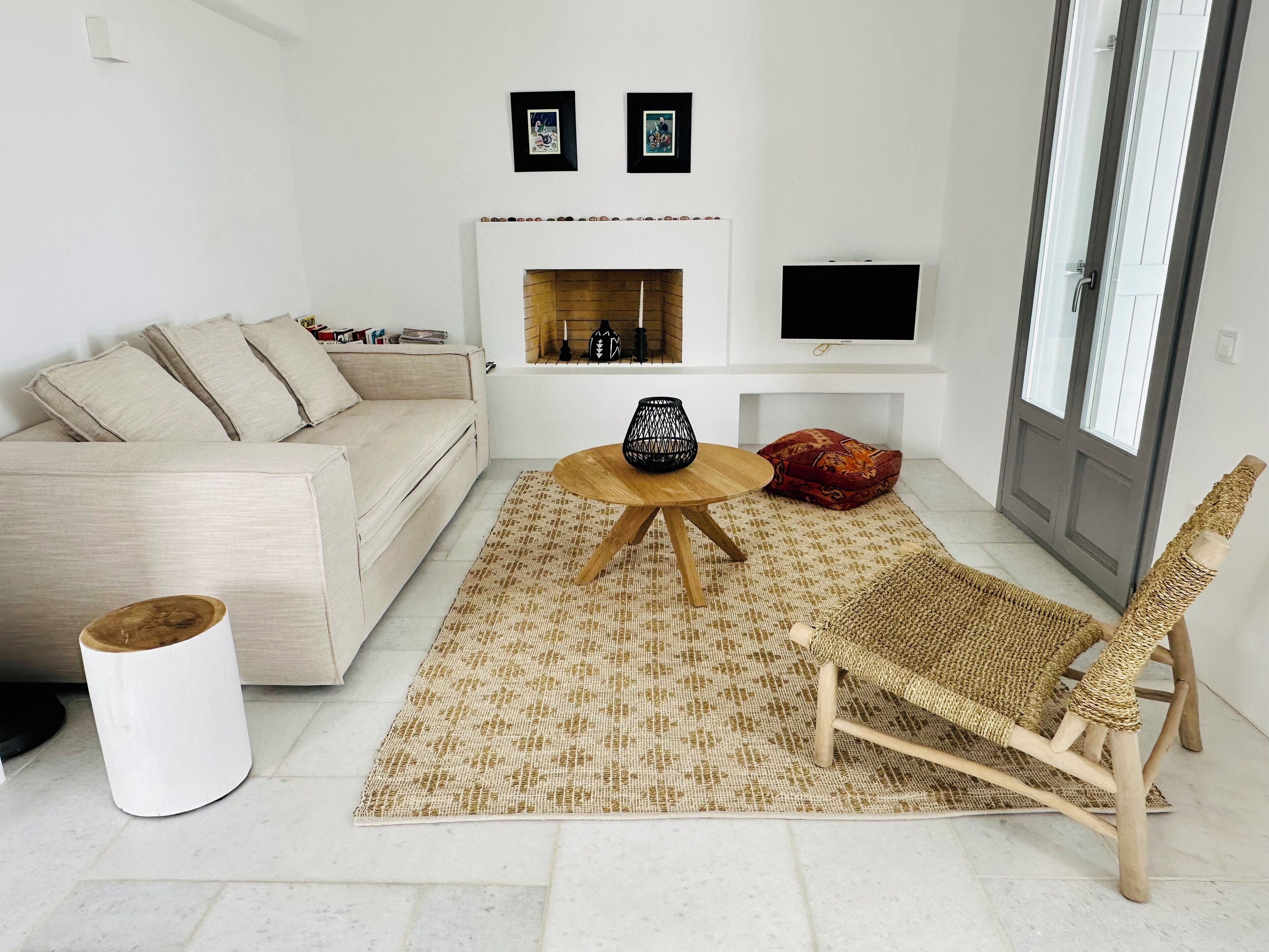 A modern living room with a sofa, a round wooden coffee table, a woven rug, a wicker chair, and a fireplace with framed pictures above it.