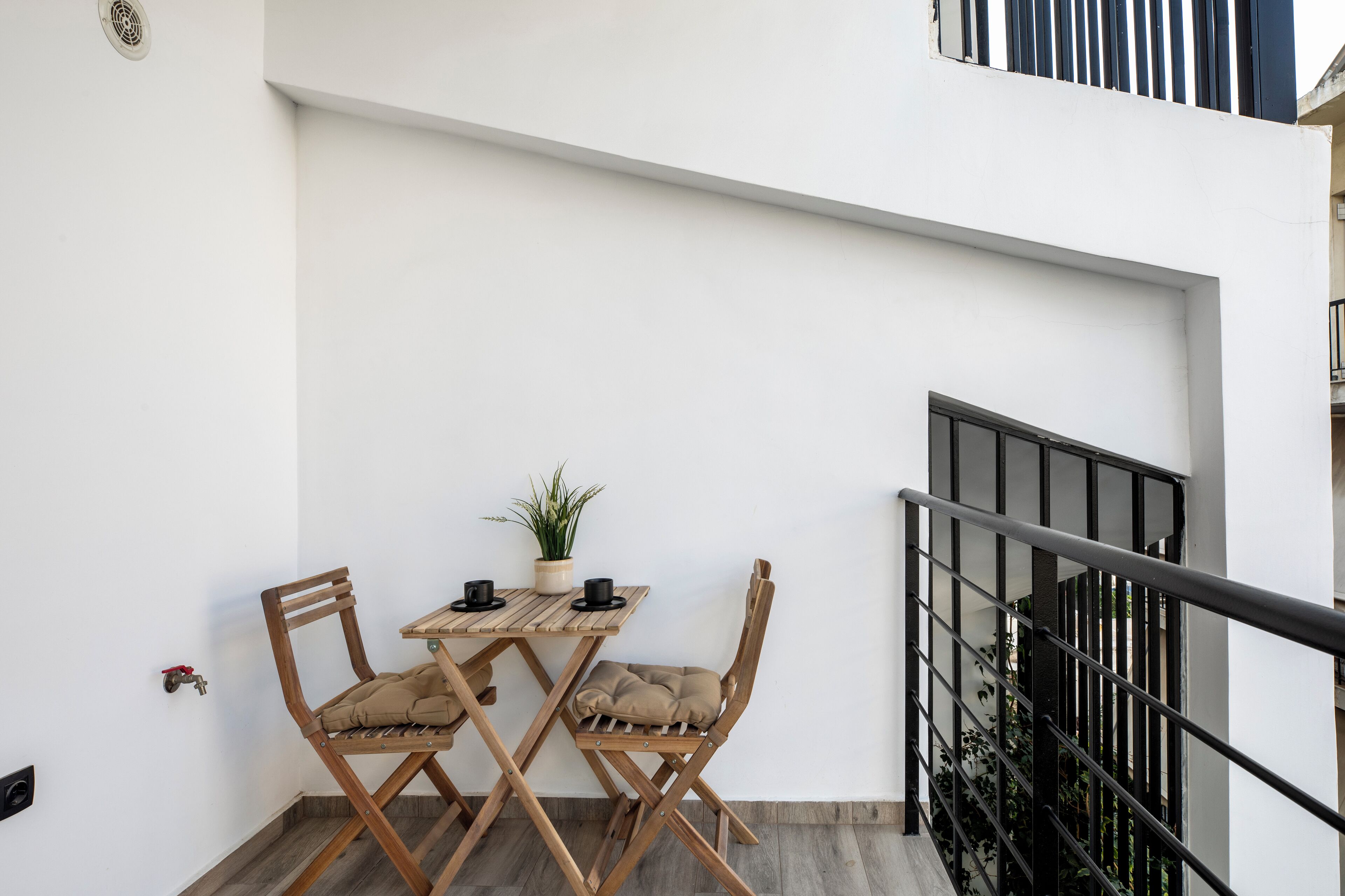Ein kleiner Balkon mit einem Holztisch, zwei Stühlen, einem Blumentopf und einem schwarzen Geländer.