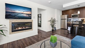 A modern living room with a fireplace, a large TV mounted on the wall, and a kitchen area with dark wood flooring and stainless steel appliances.