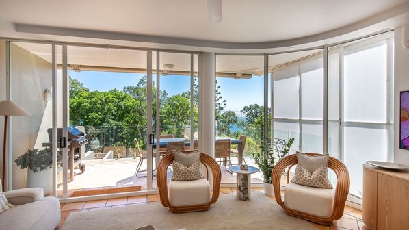 A modern living room with a curved glass wall, two cushioned chairs, a small round table, and a wooden cabinet.