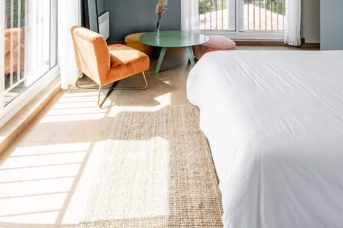 Un dormitorio moderno con una cama grande, una silla, una mesita con un florero y un balcón con cortinas.