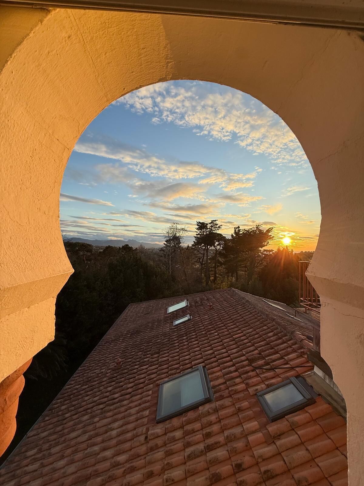 A view through an arched window of a tiled roof and a sunset.