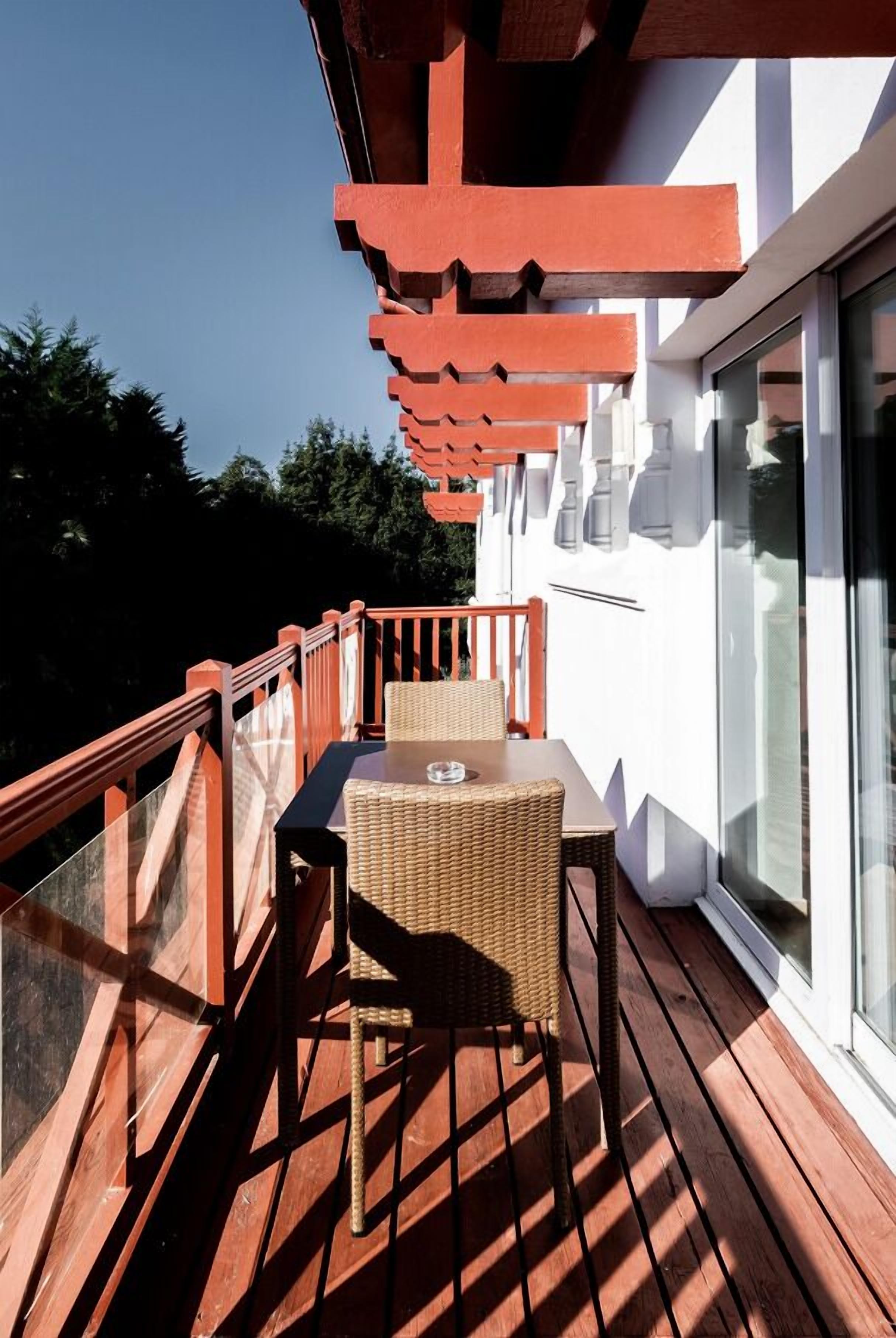 A wooden deck with a table and chairs, a glass balustrade, and a white wall with windows.