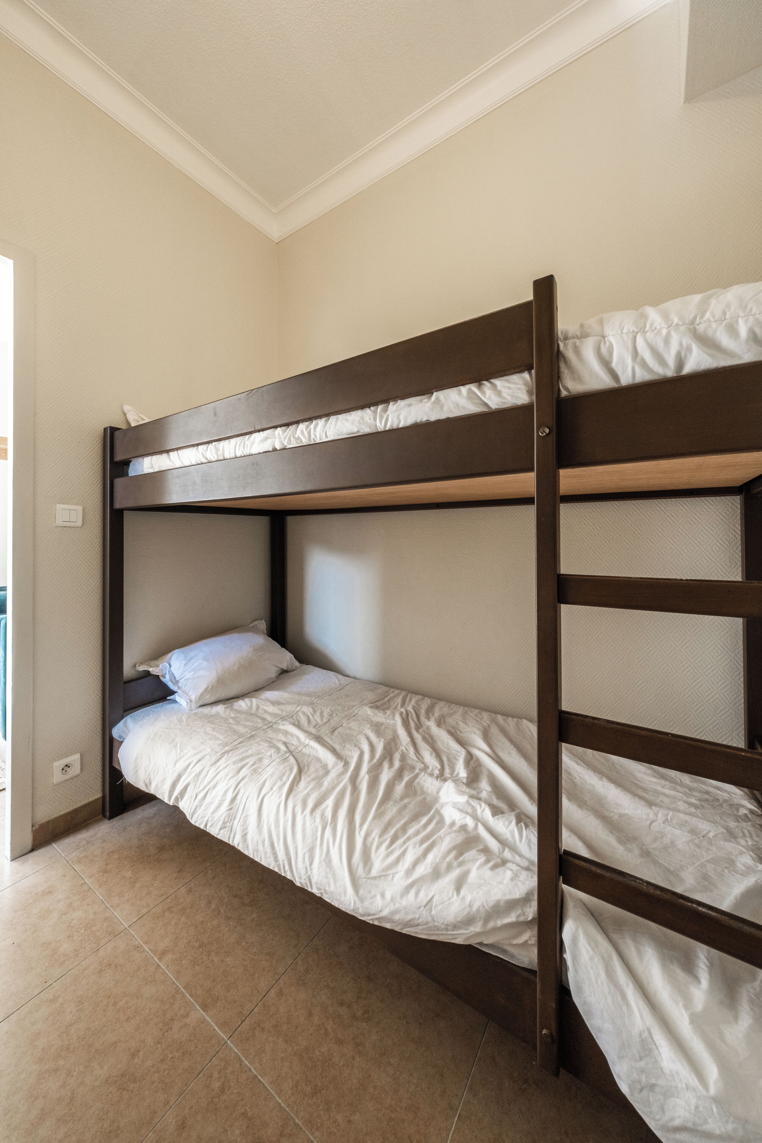 Una cama litera con estructura de madera y ropa blanca en una habitación con paredes beige y suelo de baldosas.