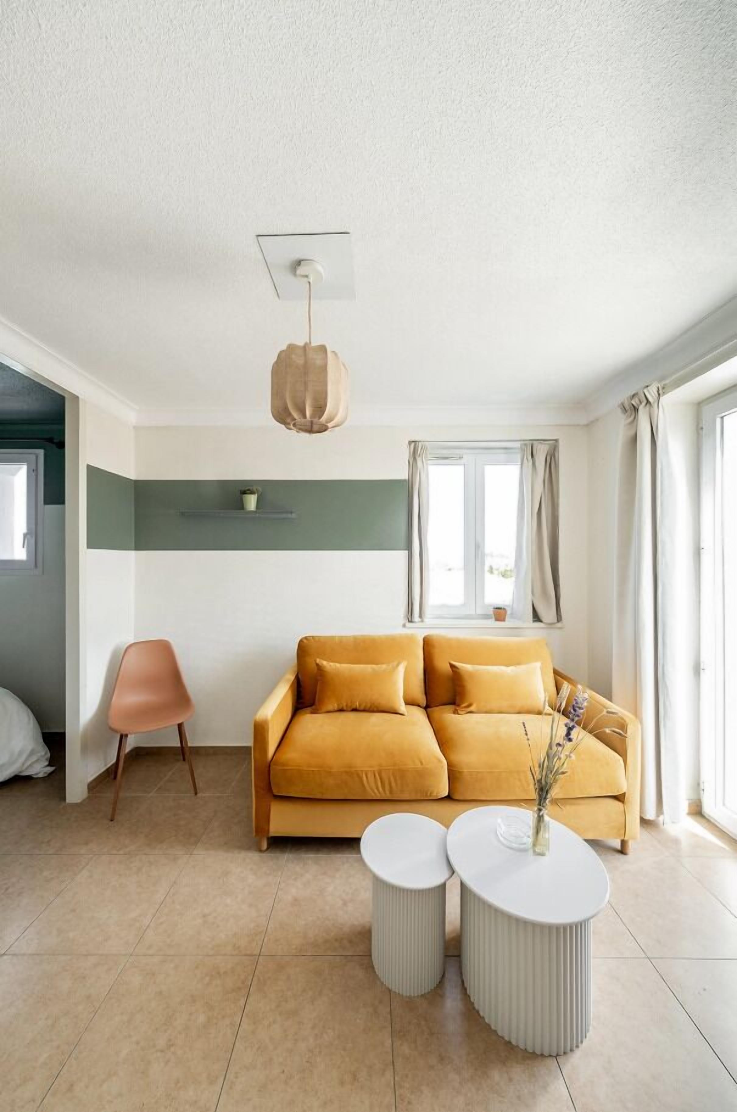 A modern living room with a mustard sofa, two white side tables, and a hanging light fixture.