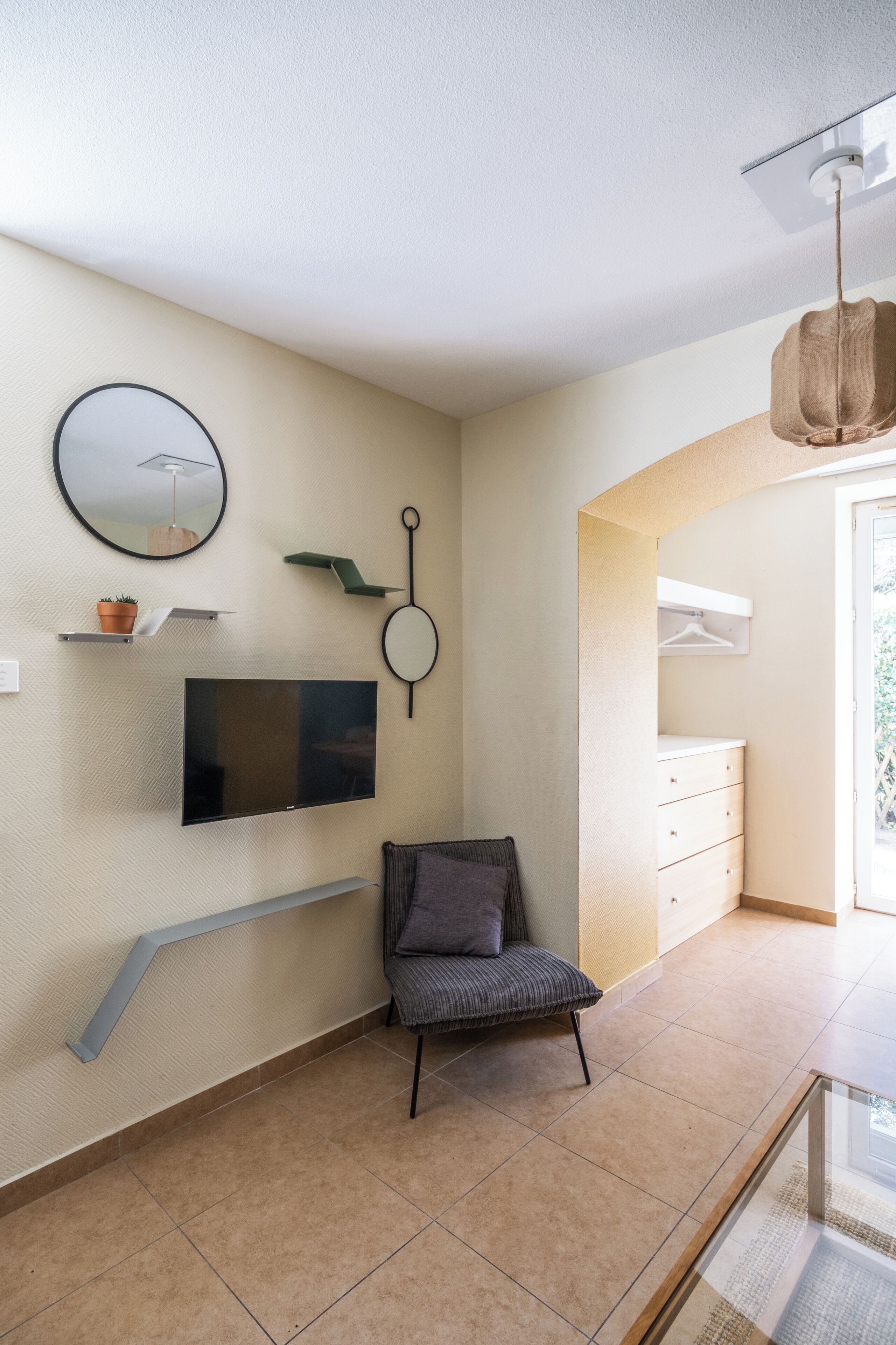 A modern room with a flat-screen TV mounted on the wall, a round mirror, a hanging light fixture, a cushioned chair, and a built-in bench.