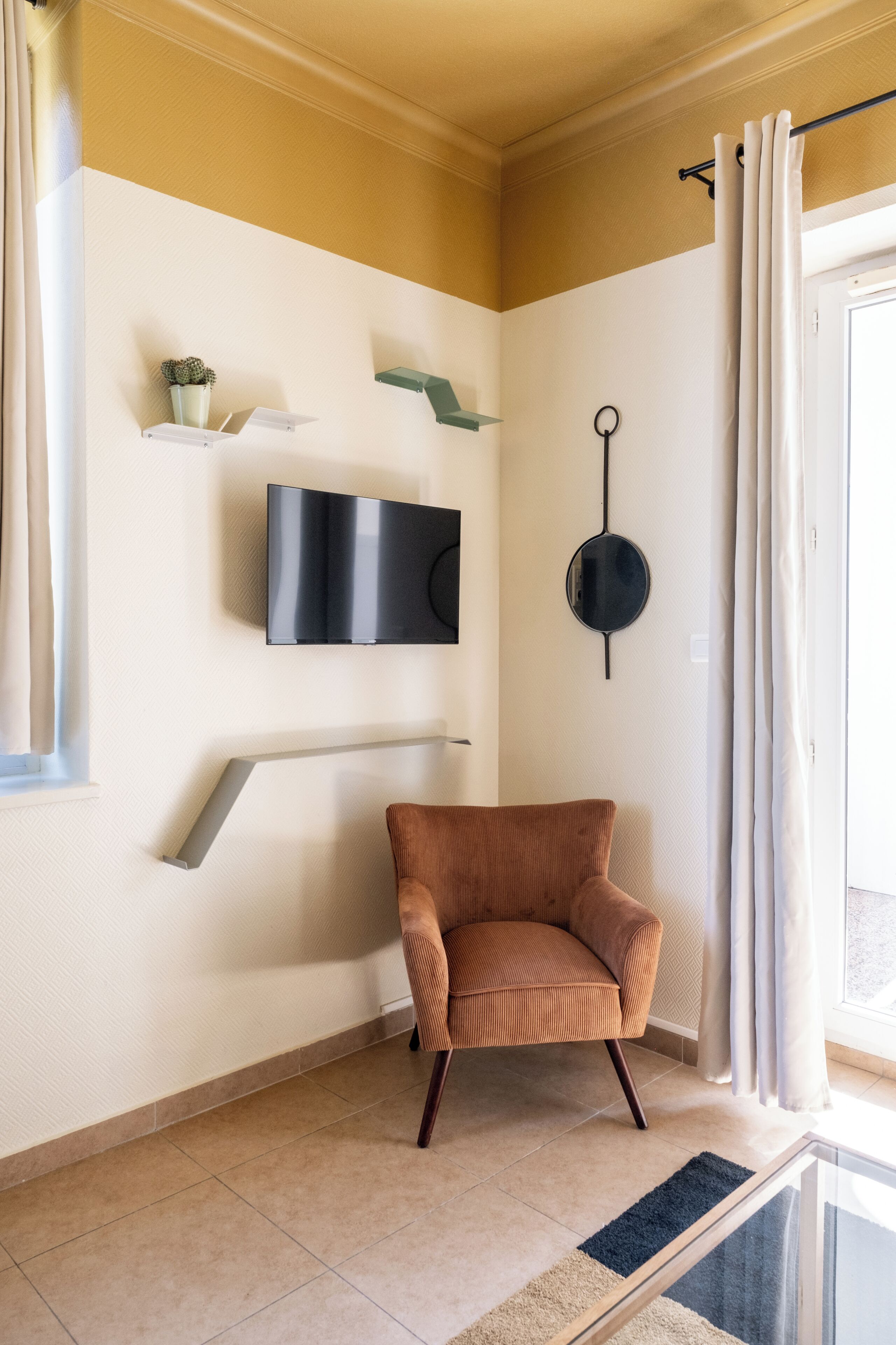 Ein Zimmer mit an der Wand montiertem Fernseher, einem Sessel, einem kleinen Regal mit einer Pflanze und einem Fenster mit Vorhängen.