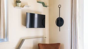 Ein Zimmer mit an der Wand montiertem Fernseher, einem Sessel, einem kleinen Regal mit einer Pflanze und einem Fenster mit Vorhängen.