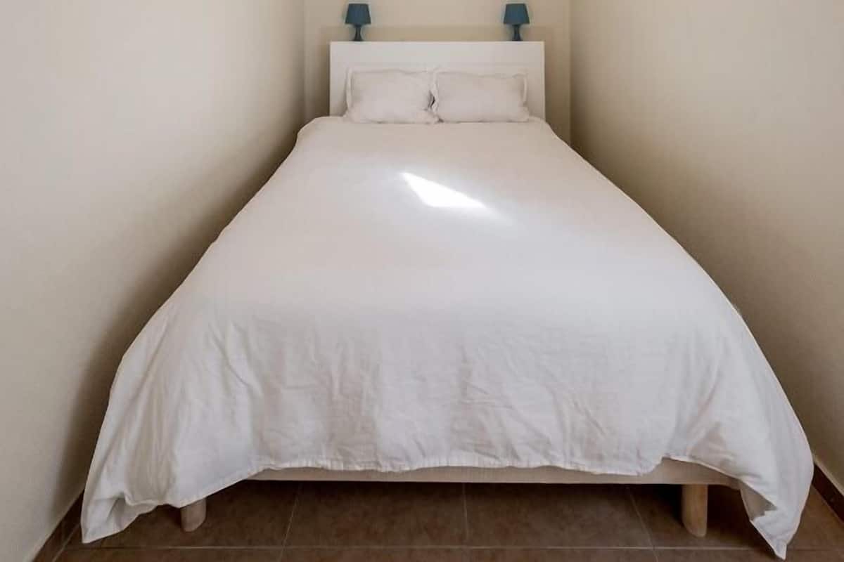 Un dormitorio pequeño y ordenado con una cama individual, dos lámparas de noche y un cabecero sencillo.