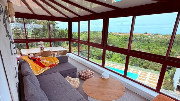 A spacious room with a large window offering a view of a swimming pool and greenery. There is a gray sofa, a wooden coffee table, and an orange sofa. - Résidence Château d'Acotz (Saint-Jean-de-Luz)