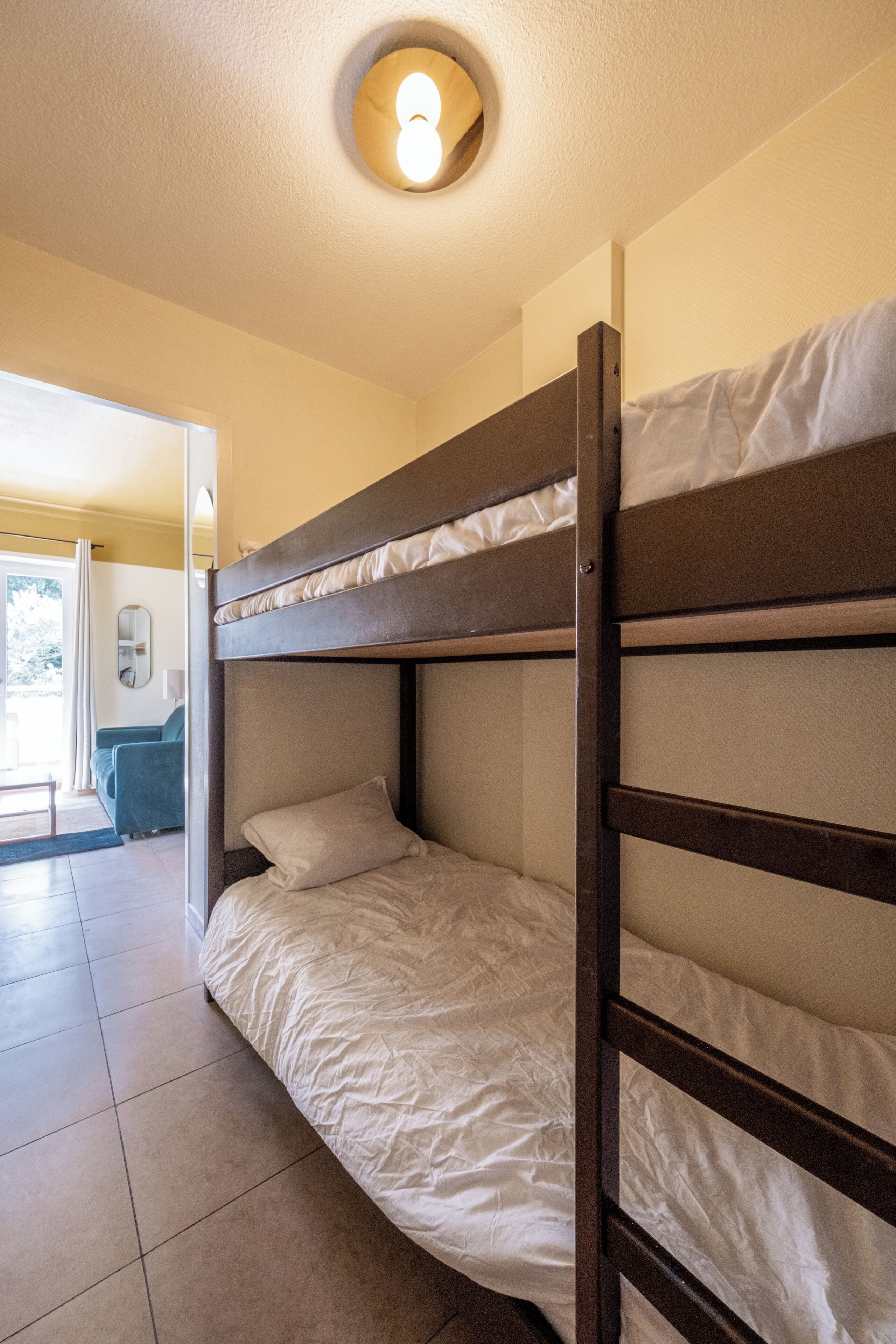 Habitación con literas, una lámpara en el techo, una puerta que da a otra habitación y piso de baldosas.