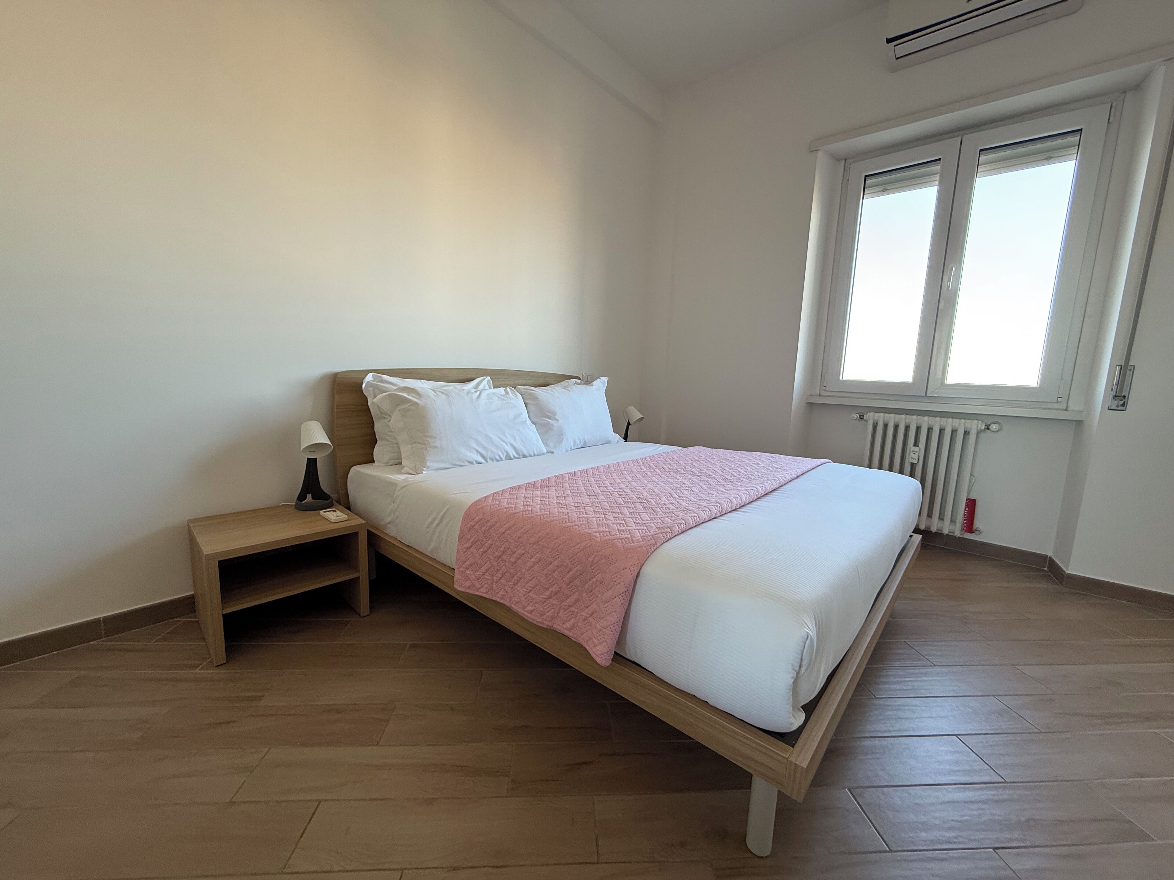 Una camera da letto con un letto in legno, biancheria bianca, una coperta rosa, un comodino con una lampada e una finestra con persiane.