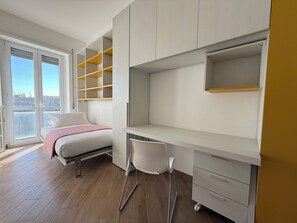 Une chambre à coucher avec un lit, un bureau et une chaise. Elle est équipée d'étagères intégrées et possède une fenêtre donnant sur des immeubles.